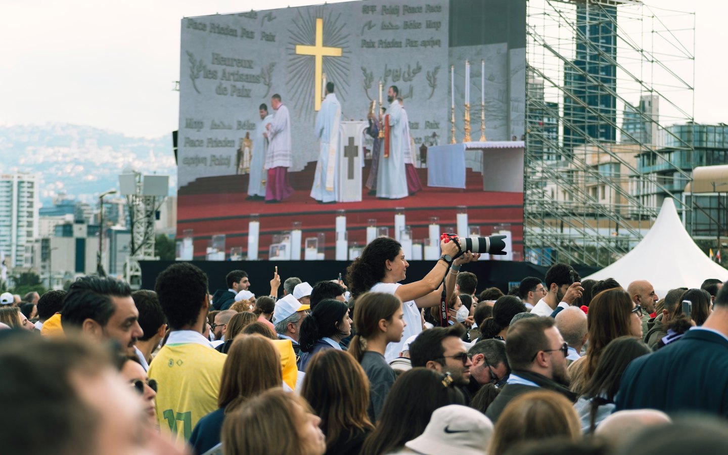 A woman photographs a religious ceremony displayed on a large screen.