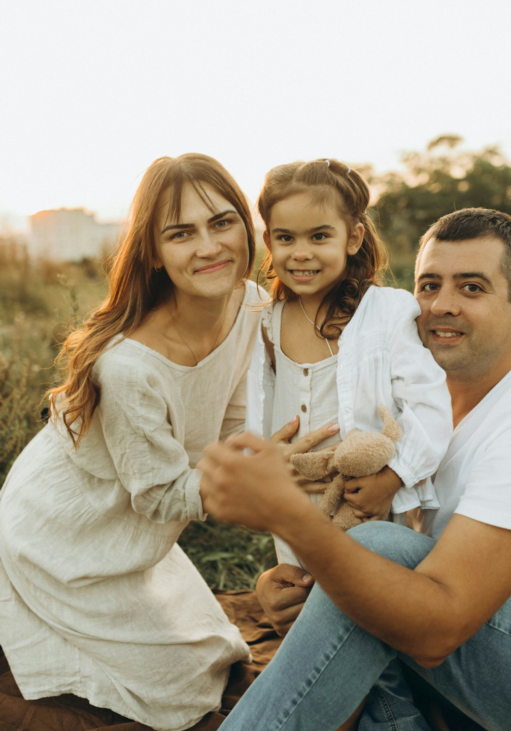 Parents and their young daughter holding a teddy bear outdoors.
